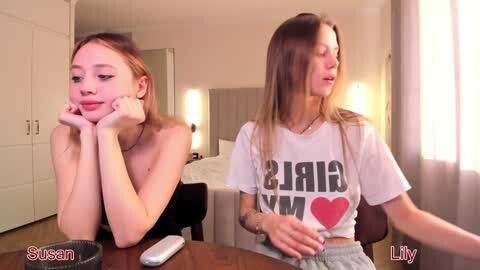 Media: Video of two young women, one with red hair, topless, the other with long brown hair, wearing a \"GIRLS\" T-shirt, sitting at a table in a dimly lit bedroom.