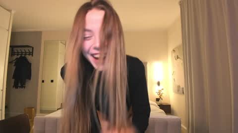Media: A video of a young woman with long, blonde hair, wearing a black top, laughing joyfully in a dimly lit bedroom with a bed, hanging clothes, and a mirror.