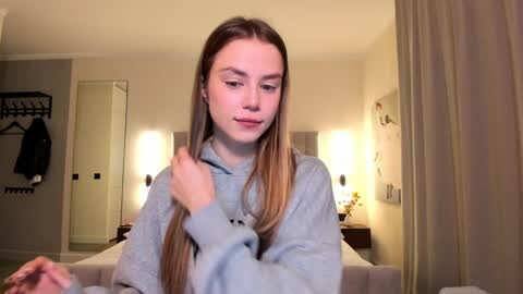 Media: Video of a young woman with long, straight, light brown hair, wearing a light gray hoodie, adjusting her hair in a modern bedroom with beige walls and a bed, lamps, and a door.