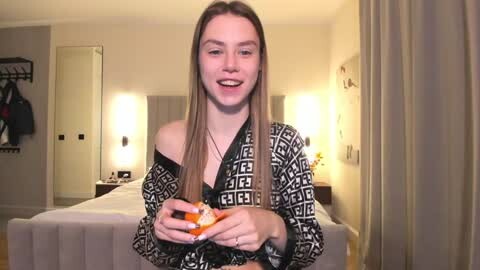 Media: Video of a smiling young woman with long blonde hair, wearing a black and white patterned robe, holding a candy bar in a cozy, warmly lit bedroom.