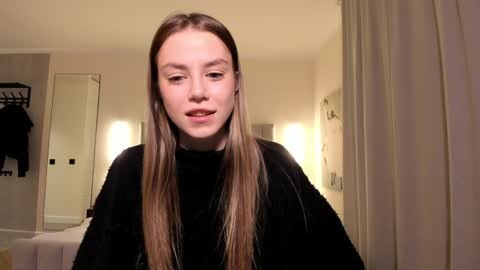 Media: Video of a young woman with straight, long brown hair, fair skin, and light makeup, wearing a black sweater, standing in a dimly lit, modern bathroom with white walls and a towel rack.