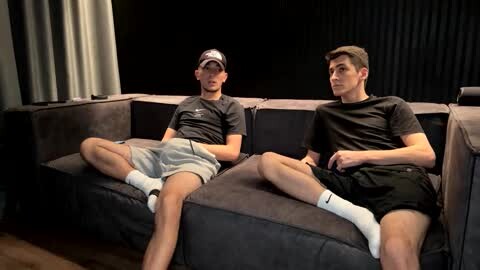 Media: Video of two young men, both wearing black t-shirts and shorts, reclining on a dark gray sectional sofa. One has a baseball cap, while the other wears white socks. Background features black curtains and a wooden floor.