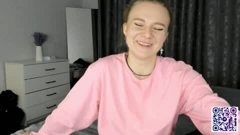Media: Video of a smiling woman with light skin and brown hair wearing a pink sweatshirt and a cross necklace, sitting on a bed in a modern, minimalistic room with gray curtains and a white dresser.