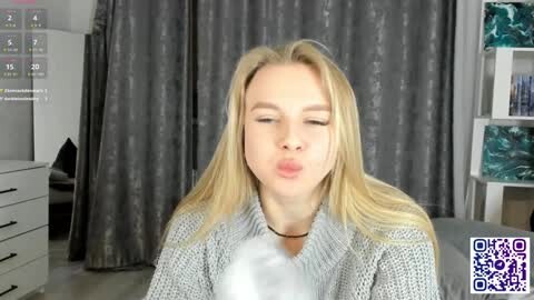 Media: A video of a fair-skinned, blonde woman with long hair, wearing a gray knitted cardigan, blowing a kiss in a modern, minimalist room with gray curtains and white furniture.