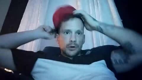 Media: A video of a man with red hair, wide eyes, and a surprised expression, holding his hair, against a white curtain background. He wears a black shirt with a white patch.