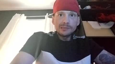 Media: Video of a Caucasian man with a mustache wearing a red baseball cap, black and white shirt, and glasses, taken indoors with a white curtain and dark walls in the background.