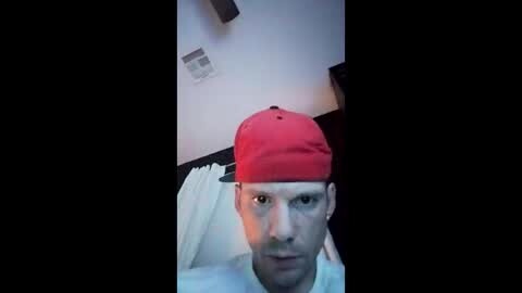 Media: Video of a man in a red baseball cap and white shirt, looking serious, taken indoors with a black background, featuring a ceiling vent and white wall.