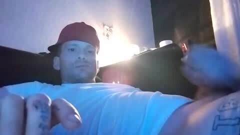 Media: Video of a muscular, shirtless man with a red hat, tattoos, and a white t-shirt, lying on a couch, illuminated by a sunlit window, blurry background.
