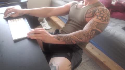 Media: Video of a tattooed man in a sleeveless gray tank top, seated at a table in a workshop, working on a wooden object with a saw.