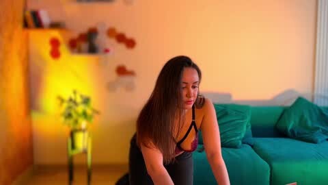 Media: Video of a woman with long, straight brown hair, wearing a black bra and panties, bending over in a cozy living room with a green couch and a lit potted plant.