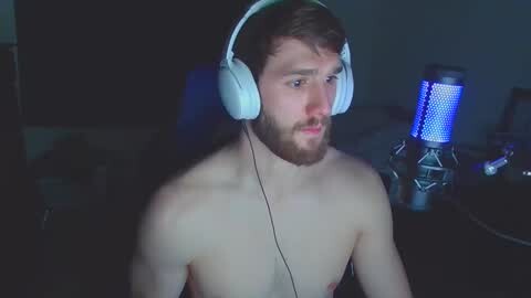 Media: Video of a fair-skinned, bearded man with short brown hair, wearing large white headphones, sitting shirtless in a dimly lit room, with a blue-lit microphone and scattered objects in the background.