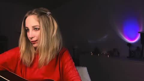 Media: A video of a blonde woman with straight hair in a red top, playing a guitar in a dimly lit room with a glowing, circular light in the background.