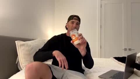 Media: Video of a young man in a black long-sleeve shirt and gray shorts, eating a sandwich on a white bed, with a laptop on the bed beside him.