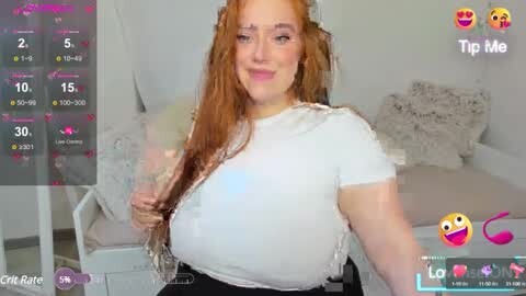Media: Video of a smiling, fair-skinned, red-haired woman with large breasts, wearing a white lace-trimmed top and black skirt, sitting on a bed in a white bedroom.