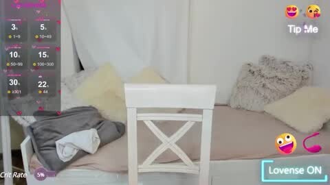 Media: A video shows a white wooden chair with a gray blanket and pillow on a beige bed with white pillows, in a cozy, minimalistic bedroom with white walls and curtains.