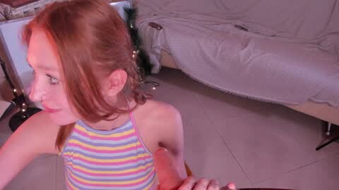 Media: Video of a young girl with red hair in a striped tank top, standing next to a bed with white sheets.