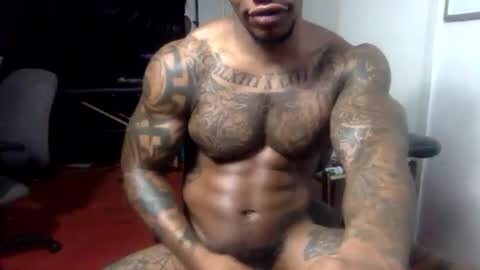 Media: Video of a muscular, tattooed Black man with a shaved head, sitting naked on a red carpeted floor. His body is adorned with intricate tattoos covering his chest, arms, and torso. The background shows a dark office with a desk and chair.