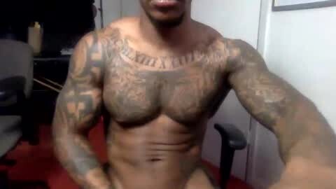 Media: Video of a muscular Black man with a large tattoo on his chest, sitting in an office chair, wearing nothing but a smile. Background shows a desk, chair, and a window.
