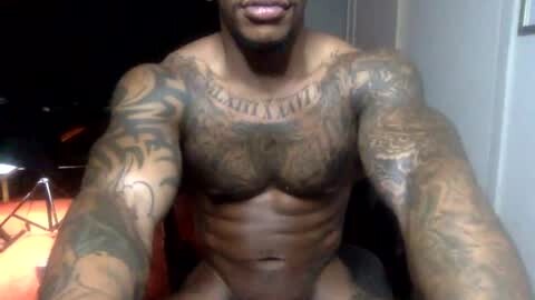 Media: Video of a shirtless, muscular Black man with numerous tattoos, including \"GOD\" on his chest, sitting in a dimly lit room. His body is well-defined, with visible abs and pectorals.