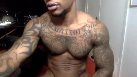 Media: Video of a muscular, shirtless Black man with extensive full-sleeve tattoos, including a large \"ROMANS\" text on his chest. He has a short beard and is sitting in a dimly-lit room with a black desk and white walls in the background.