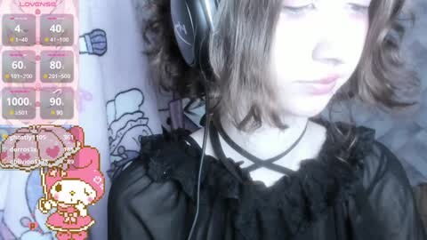Media: Video of a young woman with shoulder-length, wavy brown hair, wearing black ruffled blouse, listening to headphones, against a pastel-colored background with cartoon characters.