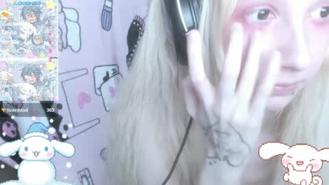 Media: A video of a pale-skinned woman with long blonde hair, wearing black headphones, holding a phone with an anime stream on it. Background has cute animal illustrations.
