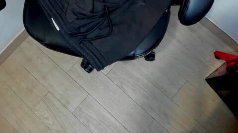 Media: A video showing a person's black backpack on a black office chair with black wheels, placed on a light wood floor with visible grain patterns. The background features a white wall and a red shoe partially visible on the right.