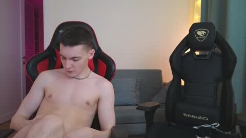 Media: Video of a young, slender, fair-skinned, and shirtless man with short brown hair, seated on a red and black gaming chair, in a dimly lit room with a grey couch and a \"Thermaltake\" gaming chair.