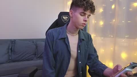 Media: Video of a young man with curly hair, wearing a dark blue shirt, playing a video game on a controller, in a modern living room with a gray couch and yellow string lights.