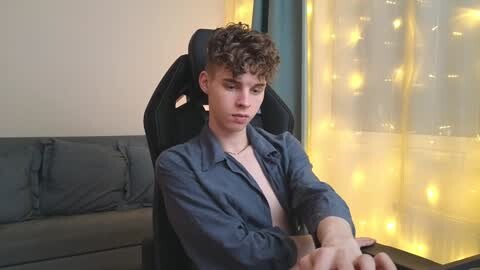 Media: Video of a young, curly-haired, fair-skinned man in a blue shirt, sitting on a black gaming chair, looking down at a tablet. Background: gray couch, yellow string lights, beige wall, window with curtains.