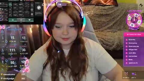 Media: Video of a young woman with long brown hair wearing a white t-shirt and colorful headphones, sitting in a cozy room with a plush bed and gaming setup.