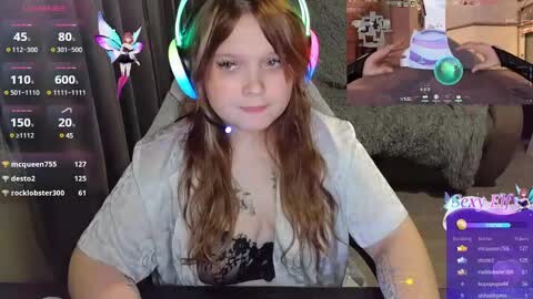 Media: A video of a young, fair-skinned woman with long, wavy brown hair, wearing a white top and headphones. She sits in a cozy, dimly lit room, surrounded by gaming stream screens showing her avatar and chat.