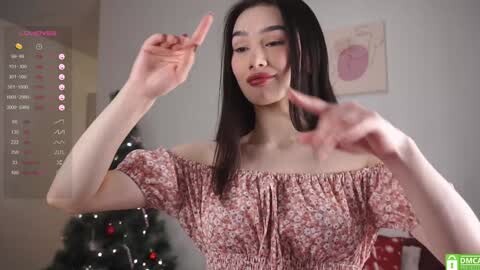 Media: Video of an Asian woman with long black hair, wearing a pink floral off-shoulder top, smiling and dancing in a brightly lit room with a Christmas tree and calendar.