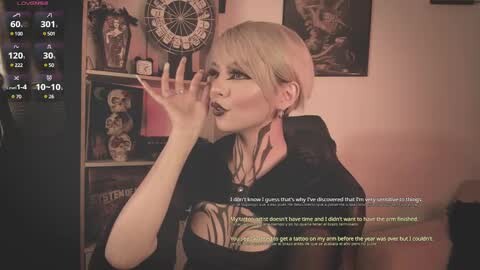 Media: Video of a pale-skinned, short-haired woman with dark lipstick, wearing a black top with a large cutout, holding a wine glass. Background includes a dartboard, shelves, and framed art. Text overlay reads, \"I don't know if I'm just being dramatic...\