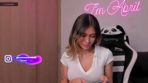 Media: Video of a young woman with long, wavy, dark hair, wearing a white V-neck top, seated in a black and white gaming chair, holding a controller, in a dimly lit room with pink neon \"I'm April\" sign.