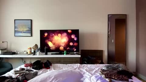 Media: Video of a cluttered bedroom with a TV displaying abstract lights on a white dresser, framed painting, open door, and messy bed.