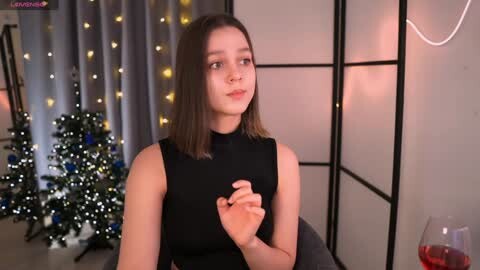 Media: Video of a young woman with straight, shoulder-length brown hair, wearing a sleeveless black dress, holding her hands together, in a festive room with a lit Christmas tree and glowing string lights.