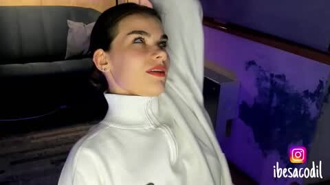 Media: A video of a young woman with light skin and dark hair in a white turtleneck sweater, stretching her arm against a purple wall with a map.