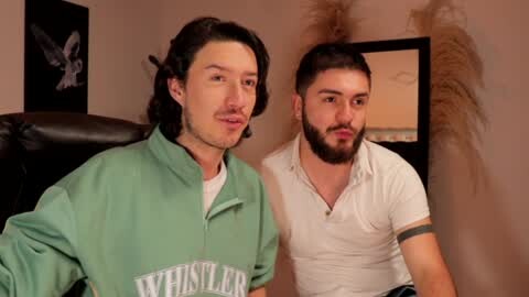 Media: Video of two men in a living room; one with shoulder-length hair, light skin, green jacket; the other with short dark hair, beard, white polo shirt.
