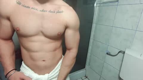 Media: Video of a shirtless, muscular man with a white towel around his waist, standing in a tiled shower with a text tattoo above his chest.