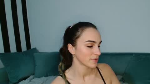Media: Video of a young woman with light skin, dark hair in a ponytail, wearing a black spaghetti strap top, sitting on a teal couch in a room with vertical black stripes on the wall.