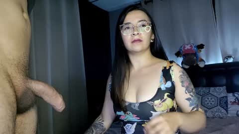 Media: Video of a tattooed woman in glasses with a floral dress, holding a man's erect penis, in a dimly lit bedroom with a bed and stuffed animals in the background.