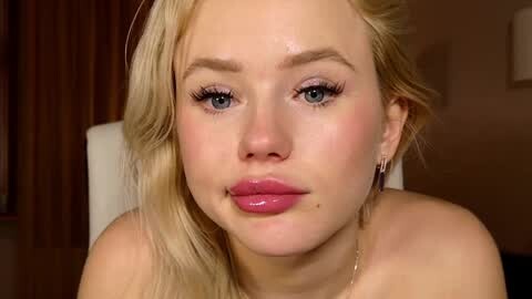 Media: Video of a fair-skinned, blonde woman with blue eyes, wearing pink lipstick and dangling earrings, with a neutral expression. Background features a wooden wall and a white chair.