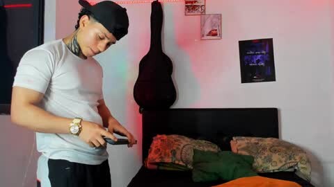 Media: Video of a fit young man in a white t-shirt, black cap, and jeans, playing a video game in a bedroom with a black couch, green and floral pillows, and posters on the white wall.