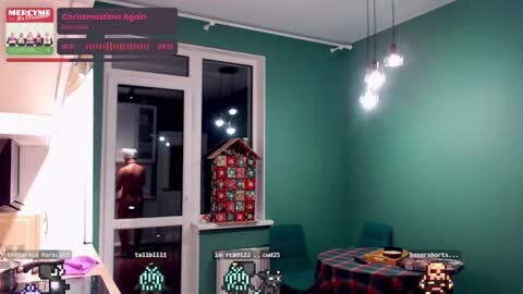 Media: Video of a teal-walled room with a man standing shirtless at a window, a decorated Christmas tree, and a dining table with a plaid tablecloth.