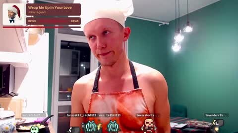 Media: A video of a young, muscular, shirtless man with fair skin and short hair, wearing a white chef's hat and an apron, in a modern kitchen with green walls.