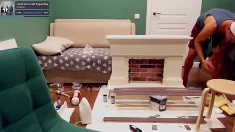 Media: Video of a messy hospital room with a green chair, white paint splattered floor, and a woman in a blue top painting a fireplace.