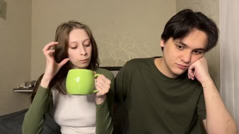 Media: Video of a young woman with long brown hair, wearing a green cardigan and a white top, holding a green cup, and a young man with short black hair, wearing a green shirt, both looking tired and sad, in a beige room with a white abstract painting on the wall.