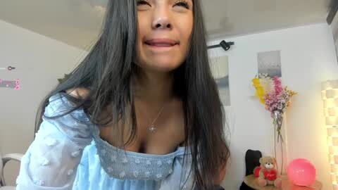 Media: Video of a young woman with long black hair, wearing a light blue off-shoulder top, smiling, in a cozy, brightly lit room with a teddy bear, flowers, and a lamp.