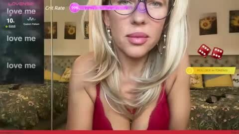 Media: A video of a blonde woman with glasses in a red bra, on a bed with floral bedding, surrounded by virtual game elements and a chat window.